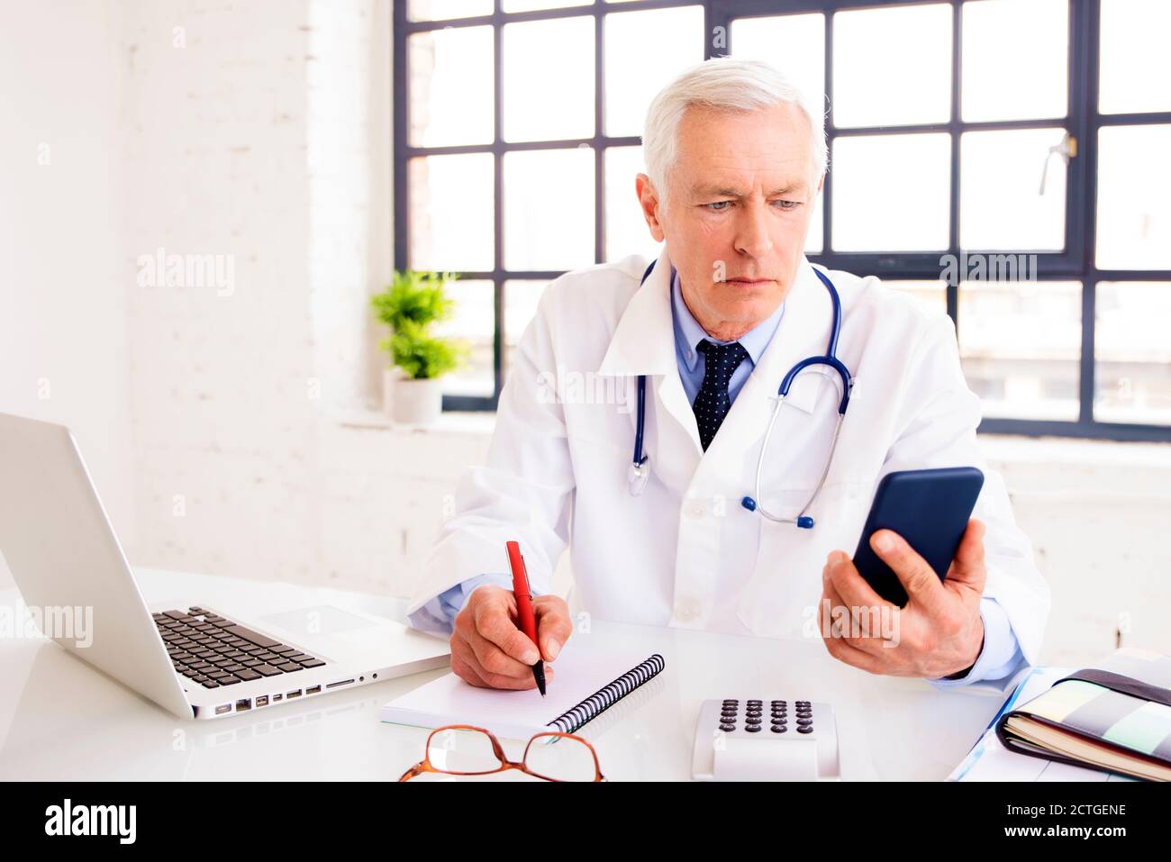 Medico maschile senior che usa il telefono cellulare e scrive qualcosa mentre si siede in ufficio del medico. Foto Stock
