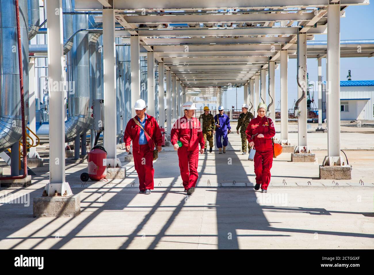 Regione di Aktobe/Kazakhstan - maggio 04 2012: Raffineria di petrolio e impianto di gas. Gruppo di lavoratori della raffineria in rosso indossare sotto le condutture camminare dritto alla macchina fotografica Foto Stock