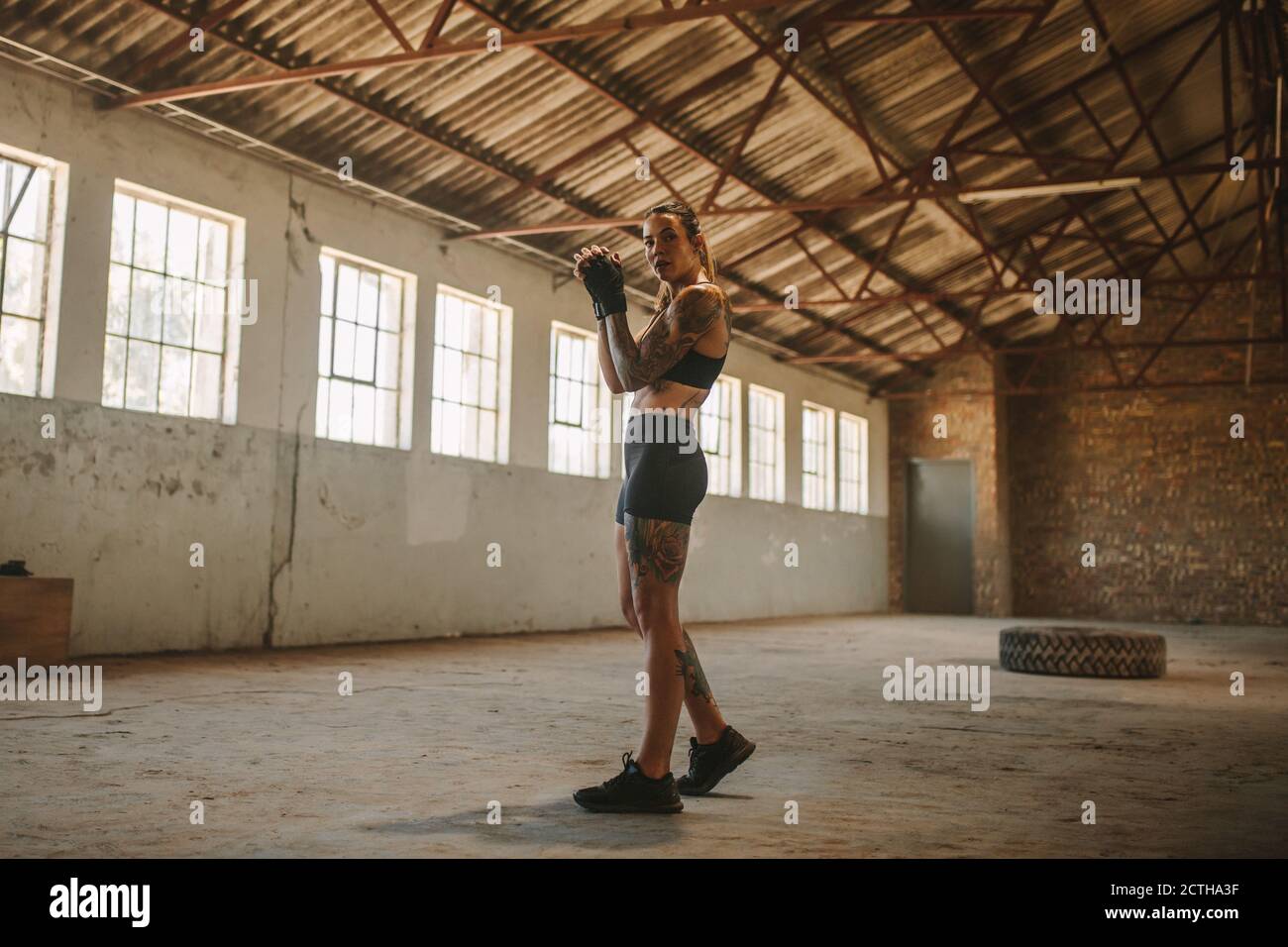 Tutta la lunghezza di una donna in abbigliamento sportivo in piedi all'interno di una vecchia ombra di fabbrica. donna fitness che pratica la boxe ombra in un magazzino vuoto. Foto Stock