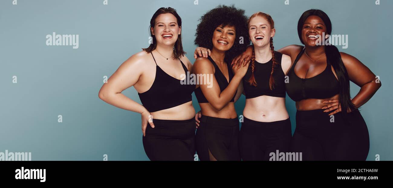 Ritratto di un gruppo di donne che presentano insieme in sportswear contro uno sfondo grigio. Multirazziale femmine con diverse dimensioni in piedi insieme alla ricerca Foto Stock