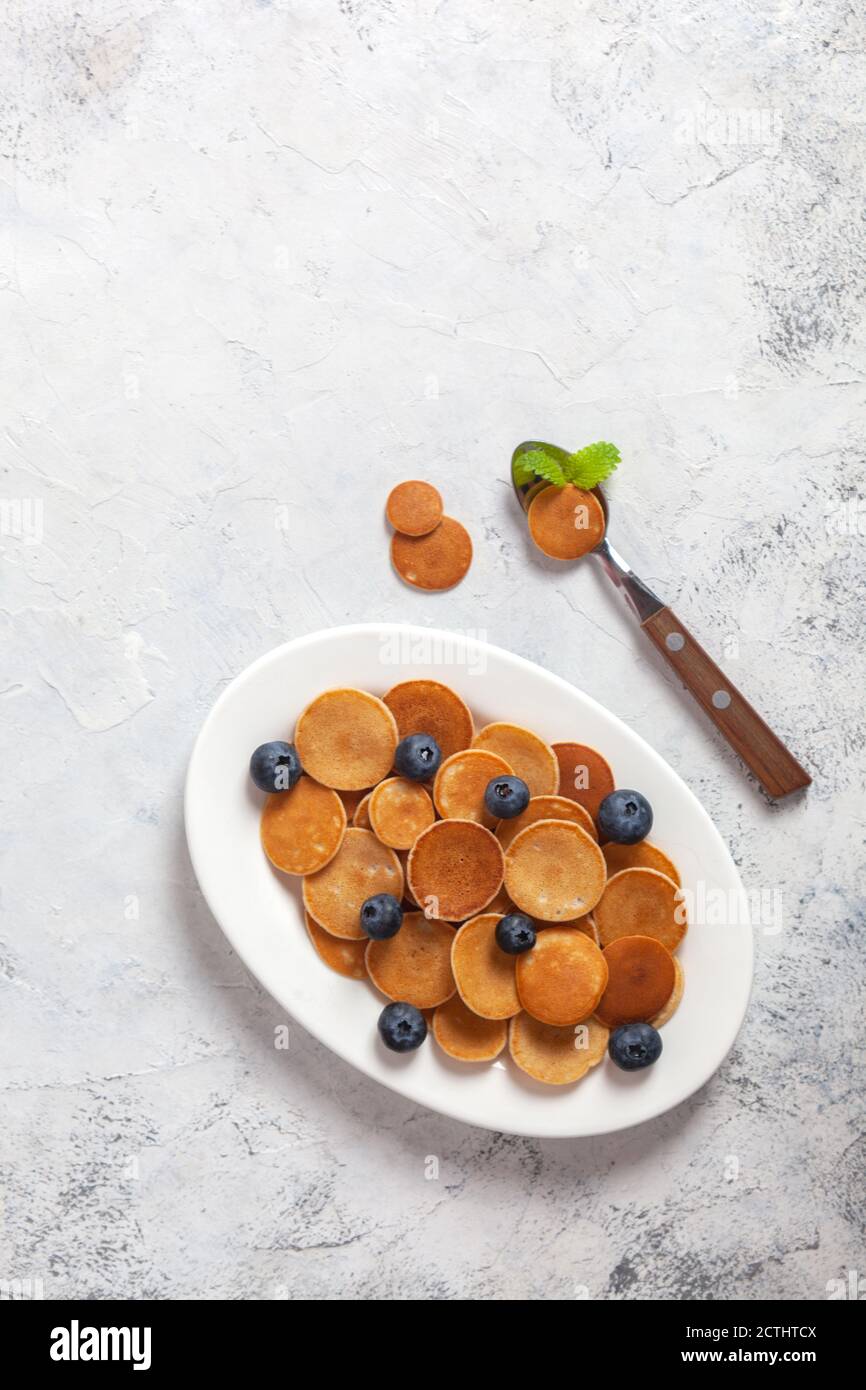 Colazione casereccia alla moda, cereali pancake, mini pancake su piatto bianco, fondo in cemento con cucchiaio e mirtillo, foglie di balsamo al limone, menta piperita Foto Stock
