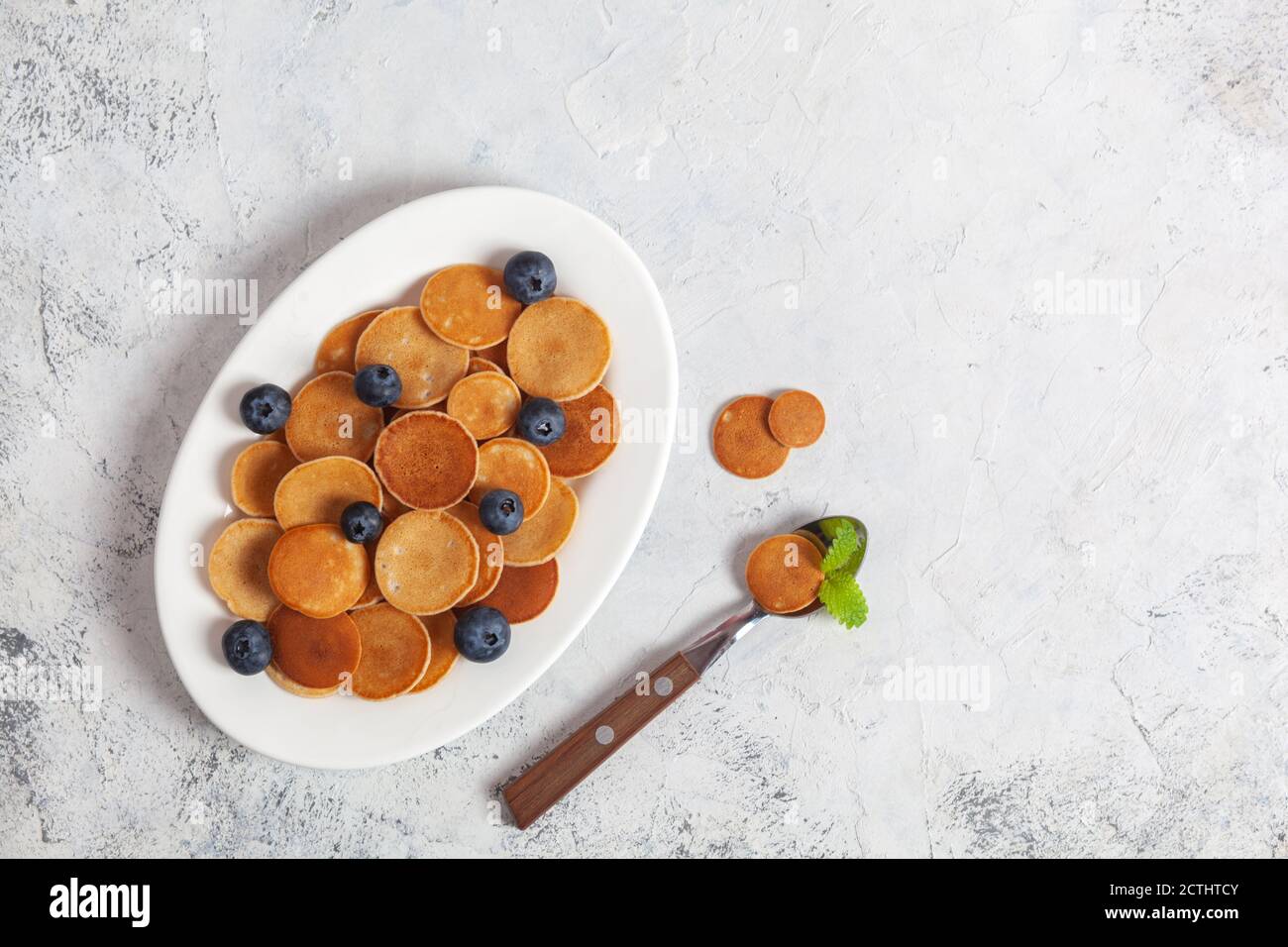 Colazione casereccia alla moda, cereali pancake, mini pancake su piatto bianco, fondo in cemento con cucchiaio e mirtillo, foglie di balsamo al limone, menta piperita Foto Stock