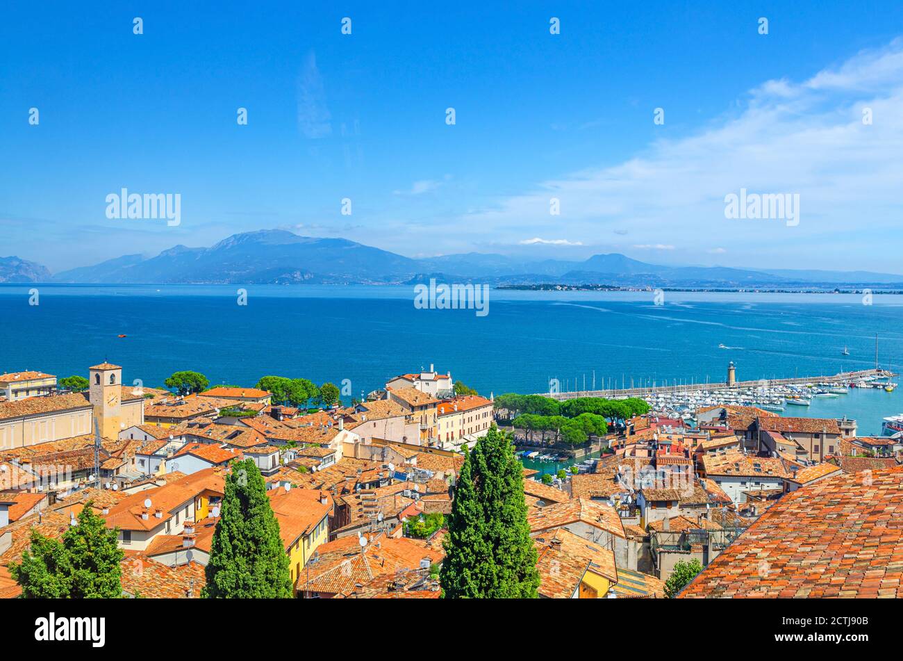 Vista panoramica aerea della città di Desenzano del Garda, tetti in tegole rosse, superficie acquatica del Lago di Garda, catena montuosa del Monte Baldo, penisola di Sirmione, faro molo, Lombardia, Italia settentrionale Foto Stock