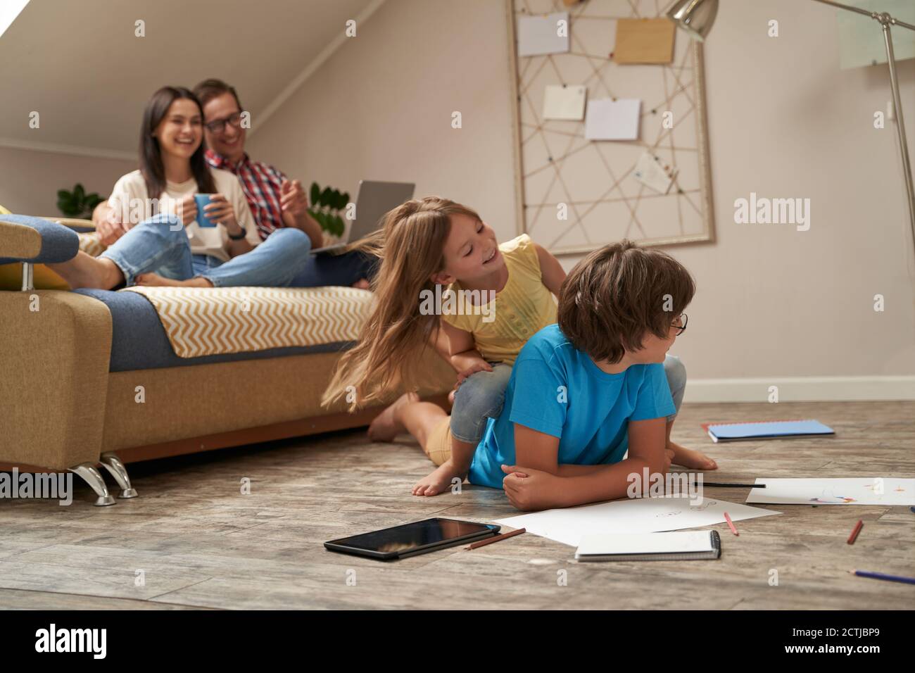 Ragazzino e ragazza che giocano insieme sul pavimento di legno in soggiorno, ridendo e divertendosi mentre i loro giovani genitori adorabili, madre felice e. Foto Stock