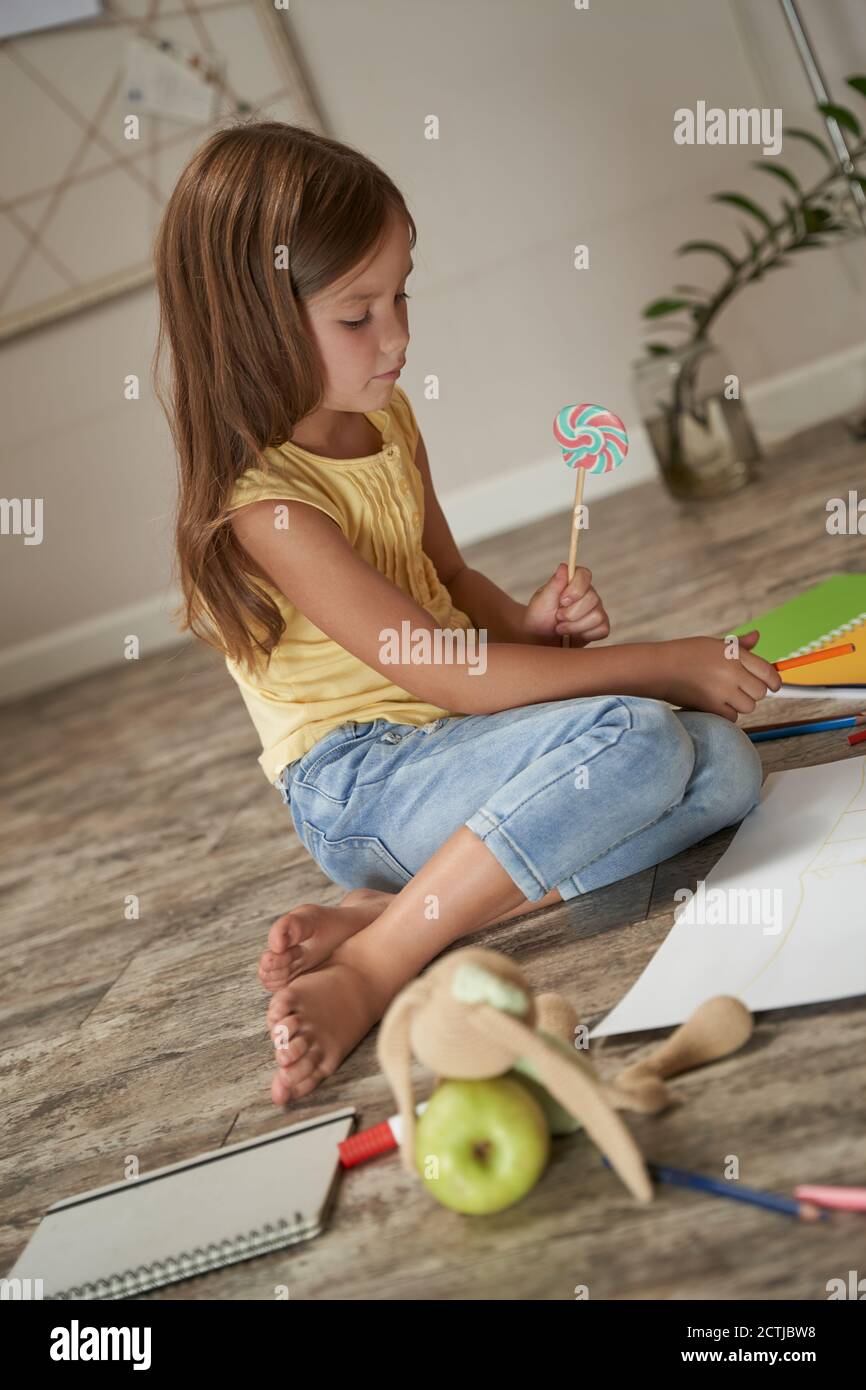 Colpo verticale di una ragazza caucasica carina tenente lollipop e disegnando con matite colorate mentre si siede sul legno pavimento nella sua stanza a casa Foto Stock