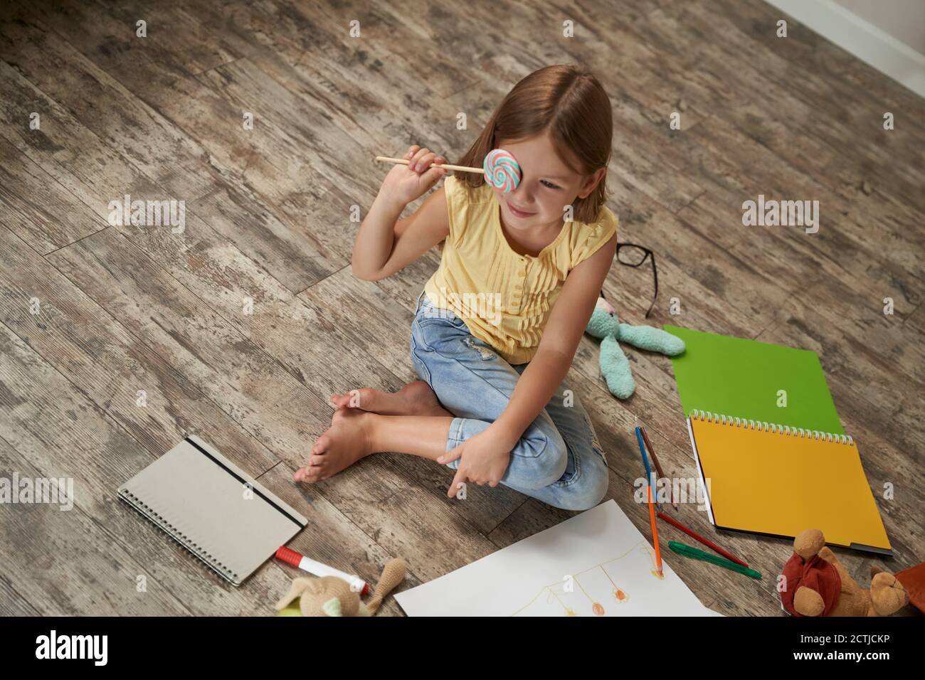 Carina ragazza caucasica tenendo lollipop e sorridente mentre si siede sul pavimento di legno nella sua stanza a casa, sta giocando con giocattoli e disegni Foto Stock