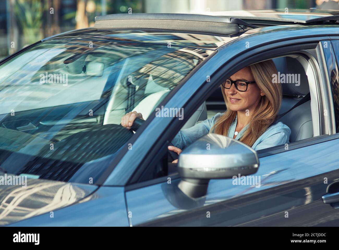Felice attraente caucasica matura donna di affari che indossa occhiali che guida la sua moderna auto attraverso la città, guardando sulla strada e sorridendo Foto Stock
