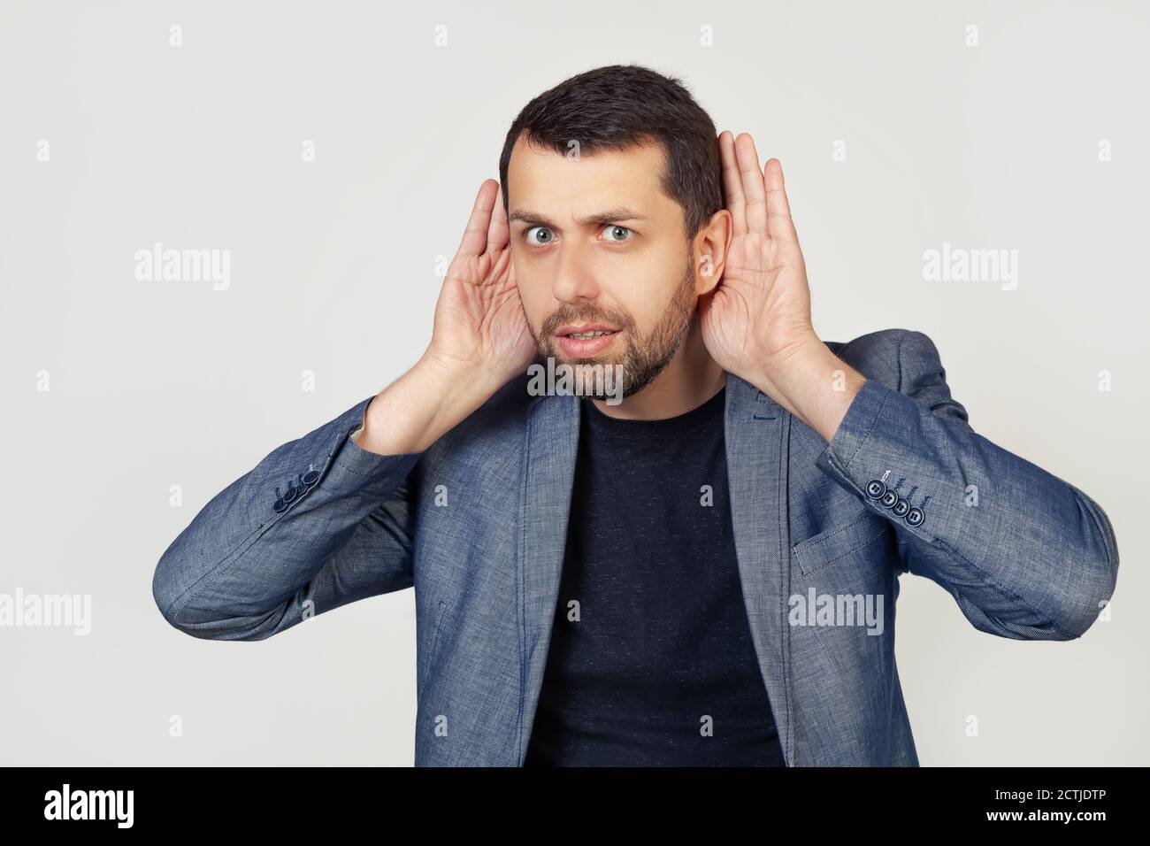 Il giovane uomo d'affari con la barba cerca di sentire entrambe le mani sull'orecchio, curioso di pettegolezzi. Problemi di udito, sordi. Ritratto di un uomo su sfondo grigio. Foto Stock