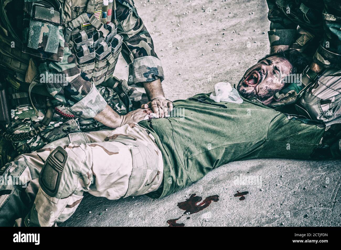 Soldati che cercano di smettere di sanguinare contro il compagno ferito che si trova a terra, sofferente e urlando a paine. Comando combattente premendo con le mani su ferita sanguinosa allo stomaco degli amici, dando assistenza di emergenza Foto Stock