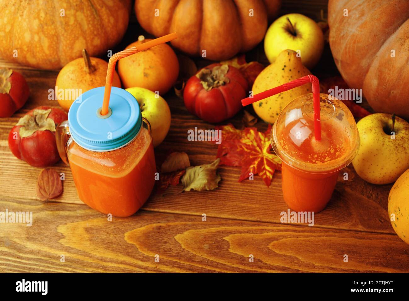 Succo di zucca e carota in tazze con tubi. Zero sprechi sull'esempio di bicchieri di vetro e di plastica. Festa della vendemmia autunnale. Zucche, persimmons, mele e foglie su sfondo ligneo. Foto Stock