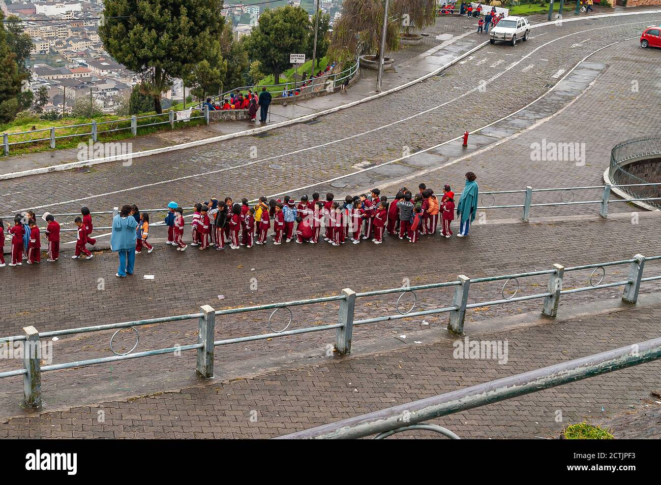 Quito, Ecuador - 2 dicembre 2008: Vista dall'alto sulla fila dei bambini della scuola in abiti maroon con insegnanti presenti. Scena di strada con verde fogliame AN Foto Stock