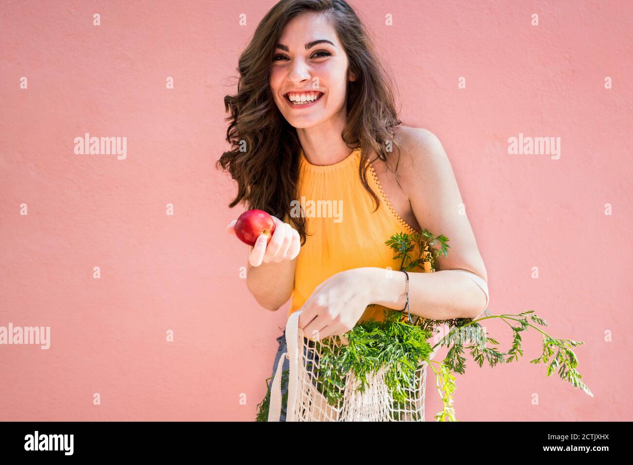 Donna allegra che tiene la mela fresca con verdure in maglia riutilizzabile sacchetto contro la parete rosa Foto Stock