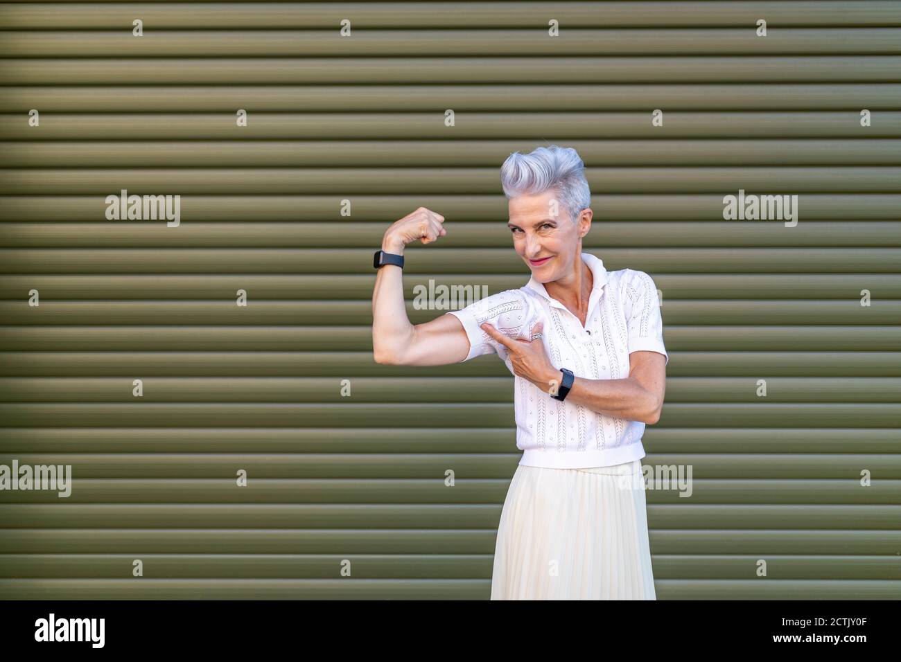 Donna anziana attiva che punta al bicep mentre flette i muscoli mentre in piedi contro l'otturatore Foto Stock Donna anziana attiva che punta al bicep mentre flette i muscoli mentre in piedi contro l'otturatore Foto Stock