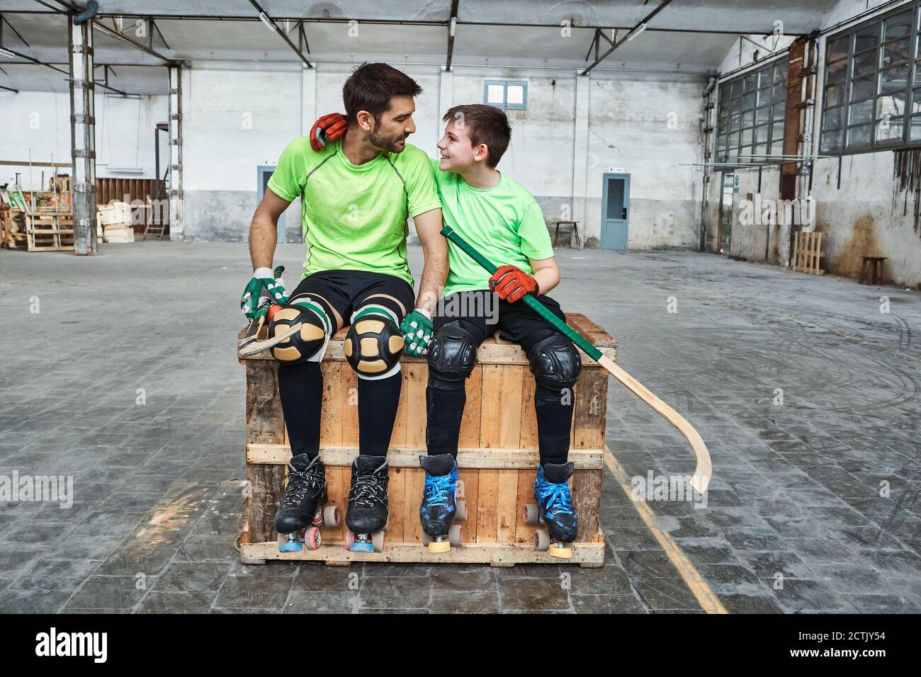 Ragazzo sorridente con braccio intorno al padre mentre si siede con l'hockey bastoni su scatola di legno a corte Foto Stock