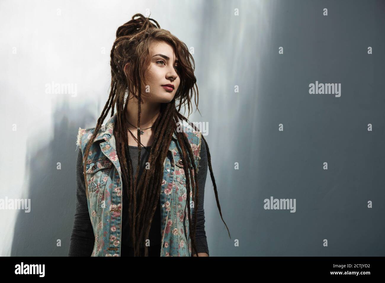 Giovane donna hipster con lunghi dreadlock in piedi contro parete grigia Foto Stock