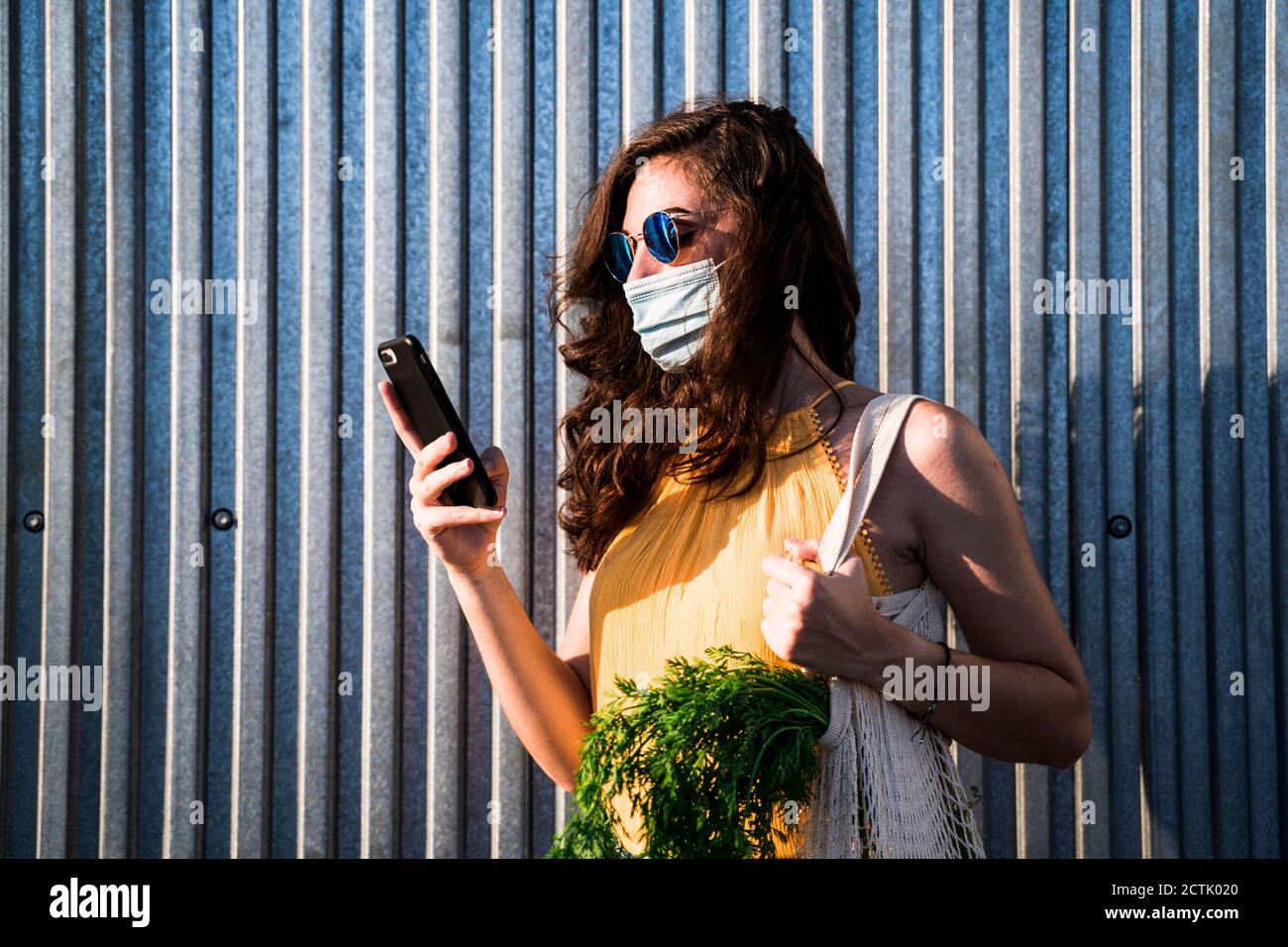 Giovane donna che indossa la maschera mentre si è in piedi con il telefono cellulare e sacchetto a rete riutilizzabile contro parete metallica Foto Stock