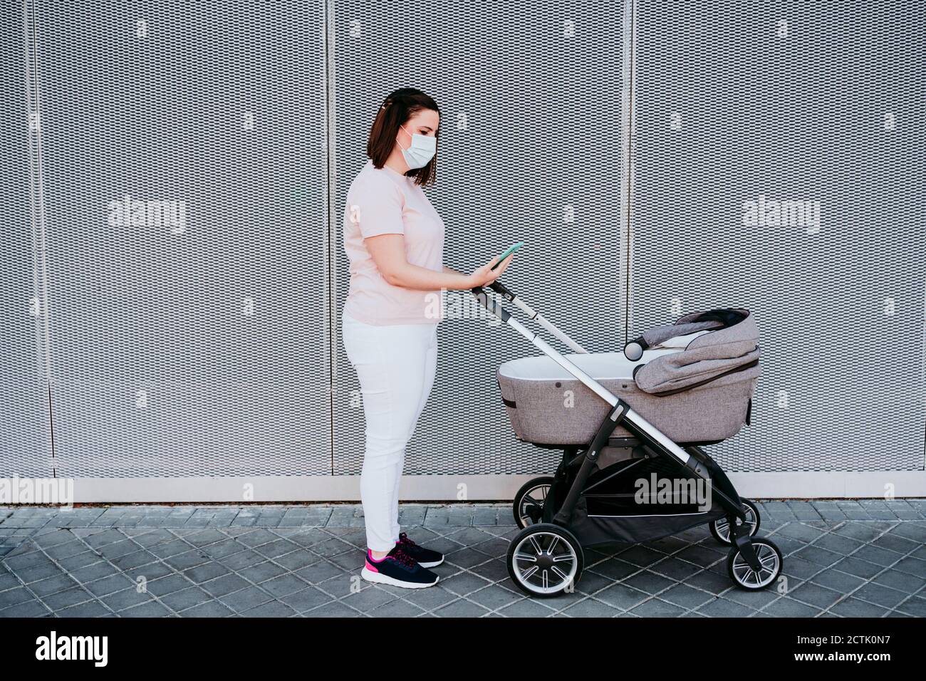 Mamma che indossa una maschera protettiva utilizzando uno smartphone in città, bambina in passeggino Foto Stock