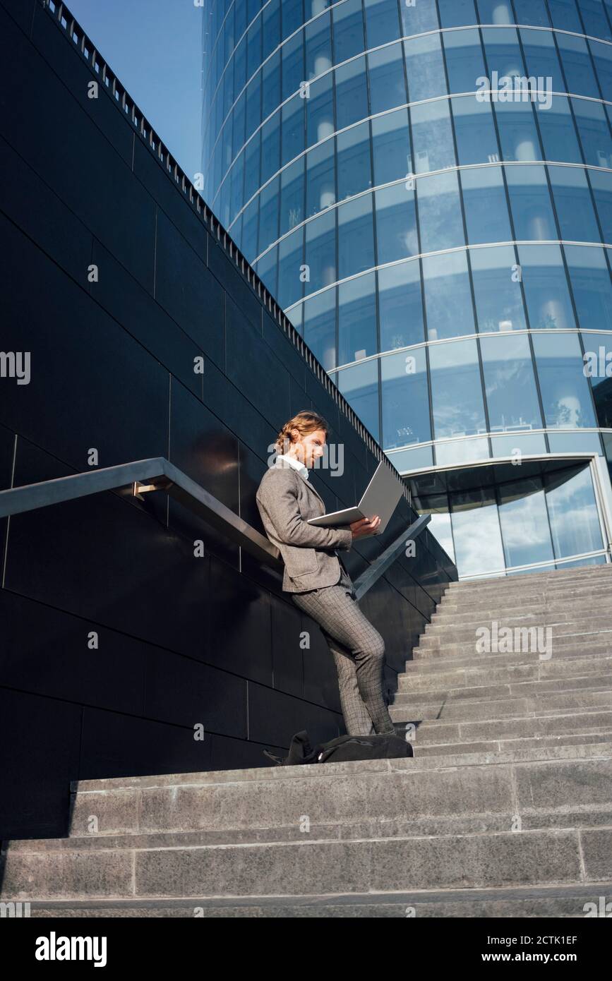 Uomo d'affari che usa il laptop mentre si appoggia contro il muro sulla scala dentro centro città durante il giorno di sole Foto Stock