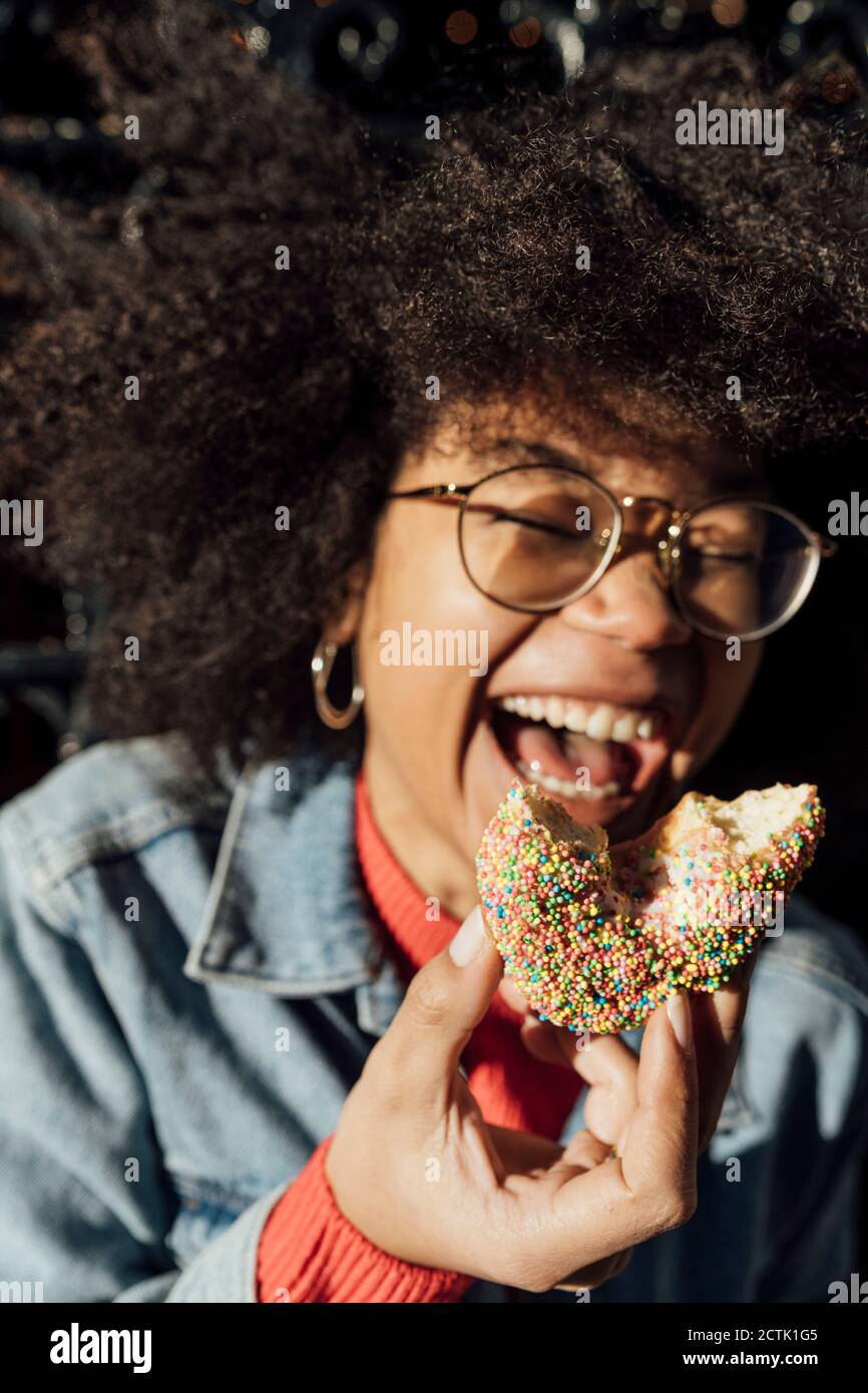 Primo piano di afro giovane donna che tiene la ciambella ridendo all'aperto durante giorno di sole Foto Stock