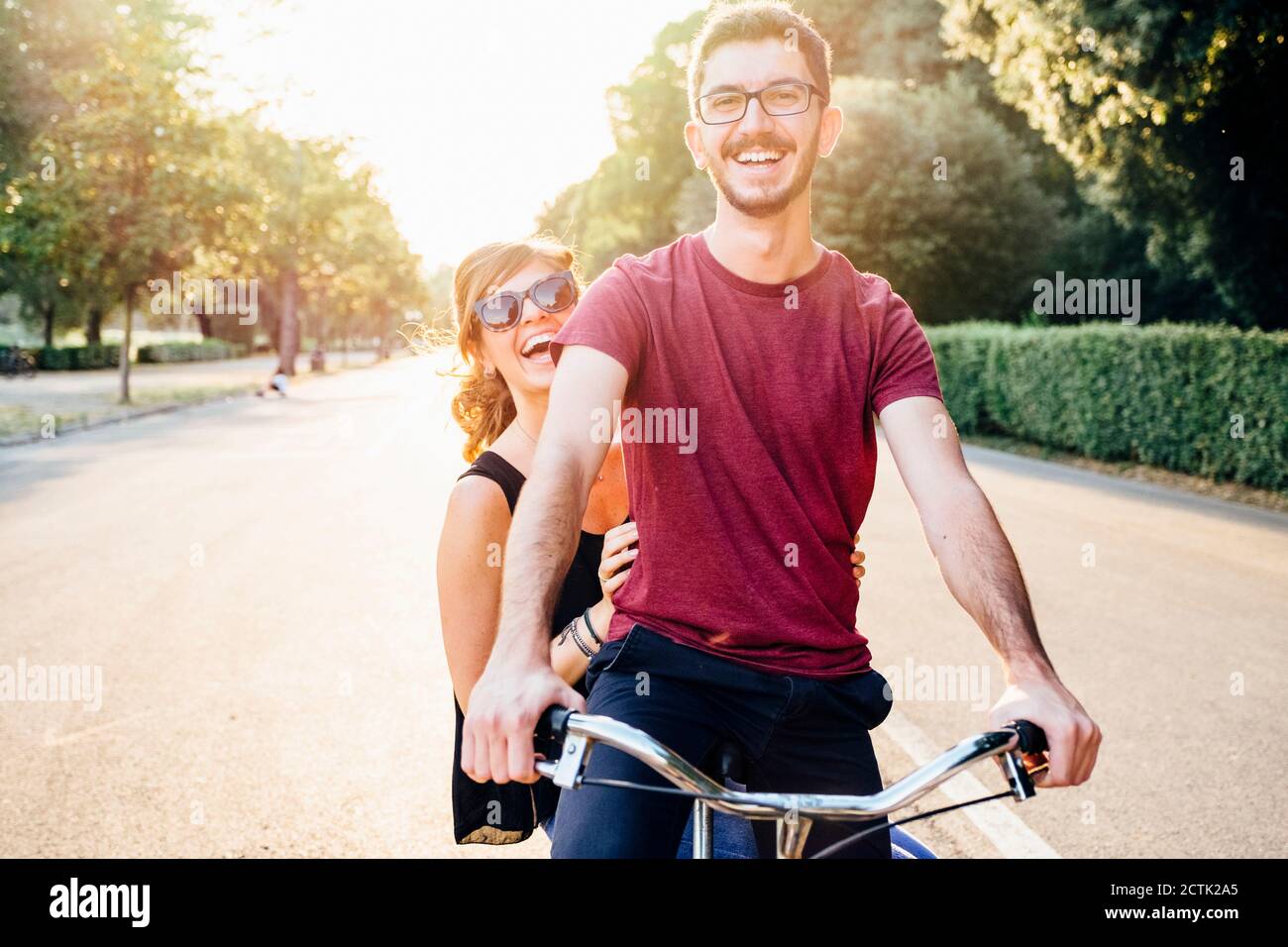 Uomo allegro con ragazza in bicicletta su strada nel parco al tramonto Foto Stock