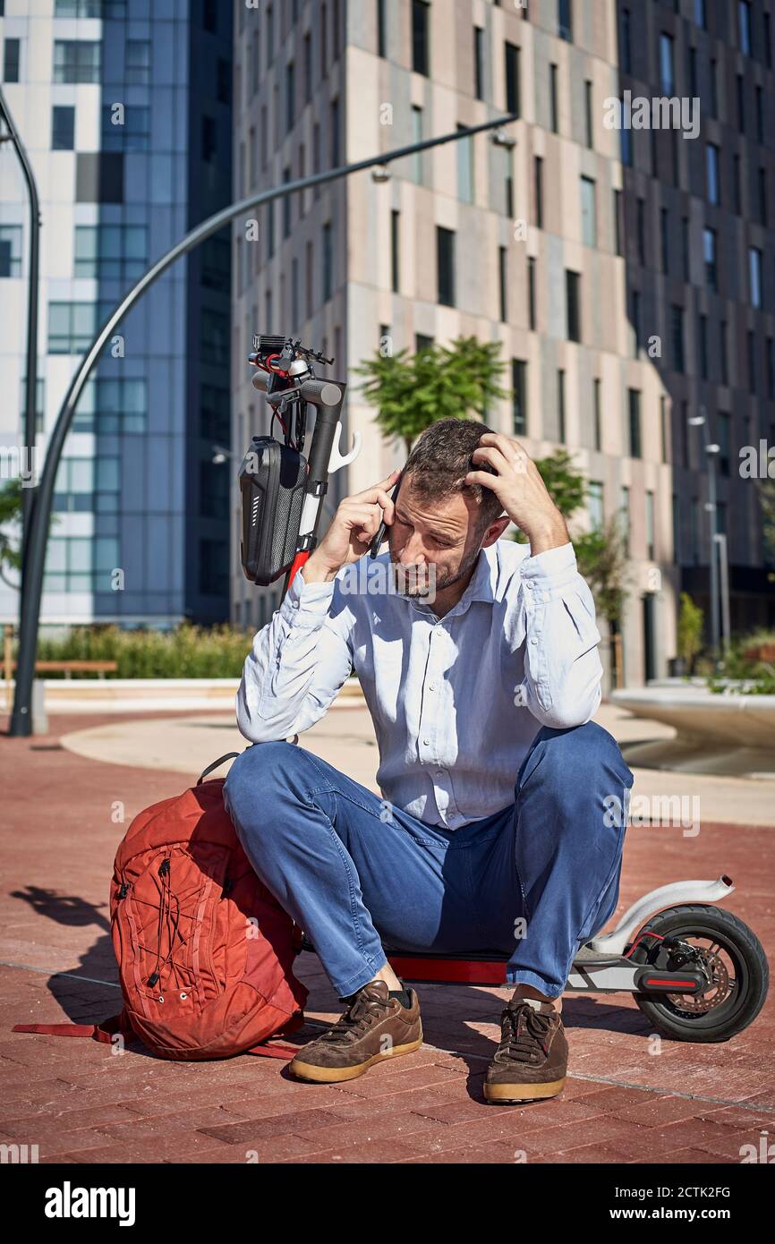 Imprenditore che parla al telefono mentre si siede su scooter elettrico dentro città Foto Stock