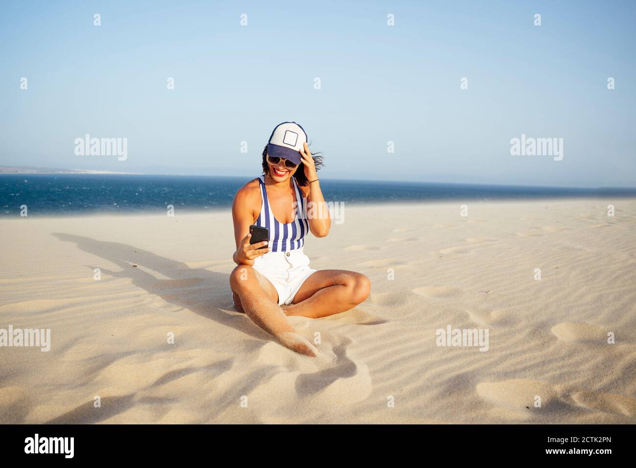 Donna sorridente che prende selfie mentre si siede sulla sabbia in spiaggia contro il cielo azzurro chiaro durante la giornata di sole Foto Stock