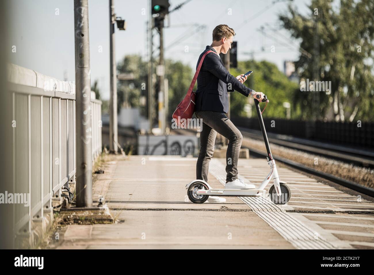 Uomo che usa lo smartphone mentre aspetta con lo scooter a spinta a. piattaforma stazione ferroviaria Foto Stock
