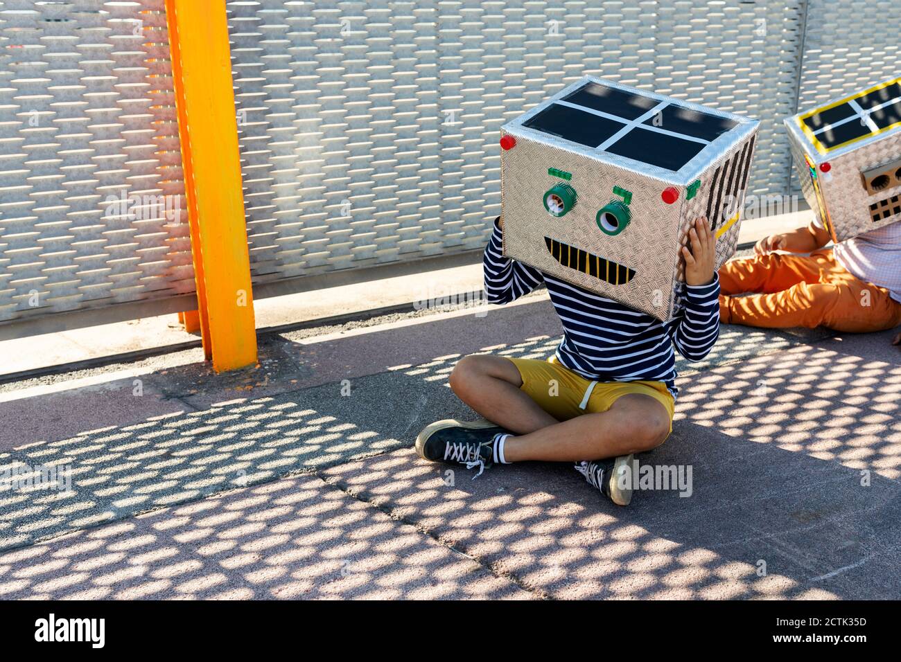 Ragazzi che indossano maschere robot fatte di scatole che si siedono su strada da recinto in città Foto Stock