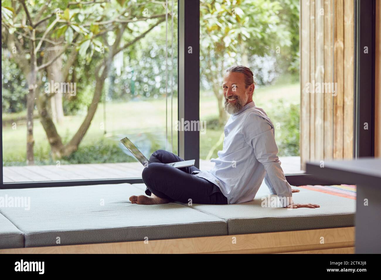 Sorridente uomo maturo con il portatore che usa il computer portatile mentre si siede a letto dalla finestra a casa Foto Stock