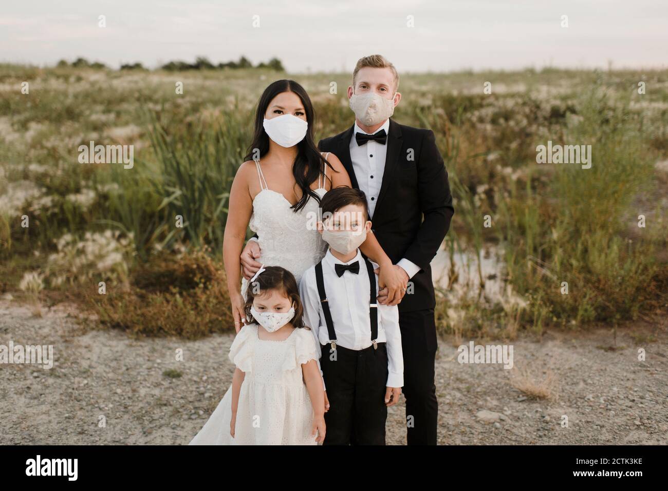 Genitori e bambini in abito da sposa con maschera mentre In piedi sul campo durante COVID-19 Foto Stock