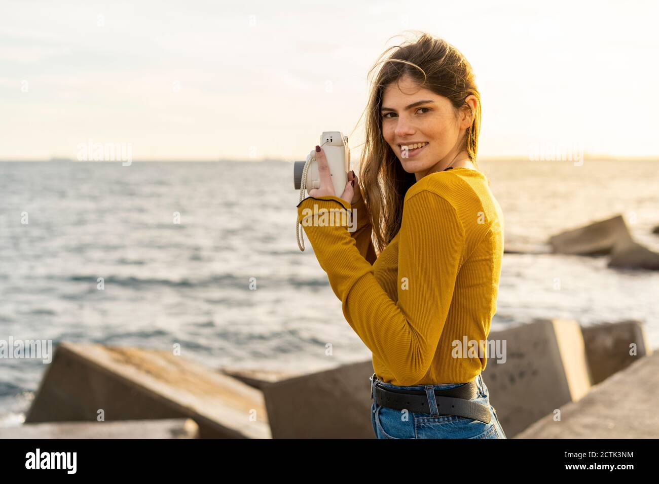 Giovane donna sorridente con macchina fotografica in piedi contro il mare Foto Stock