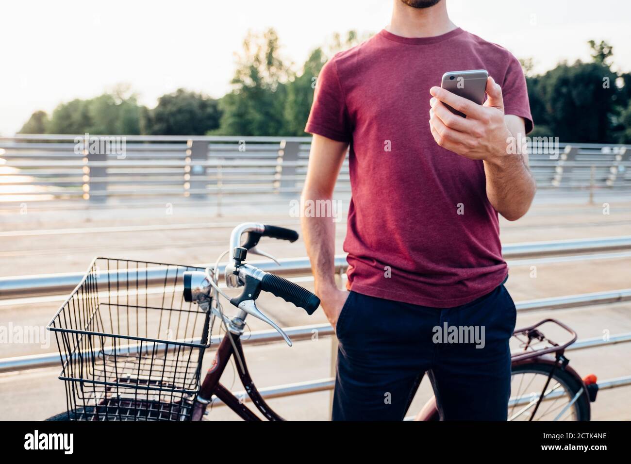 Primo piano di un uomo che utilizza lo smartphone mentre si è in piedi con la bicicletta sul ponte al tramonto Foto Stock
