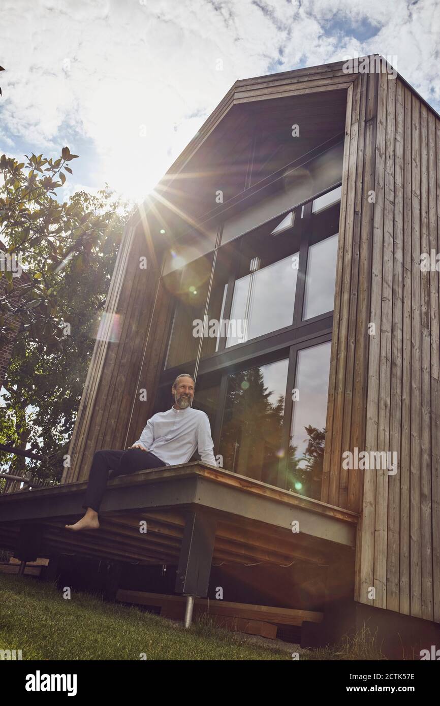 Uomo maturo seduto fuori casa piccola durante il giorno di sole Foto Stock