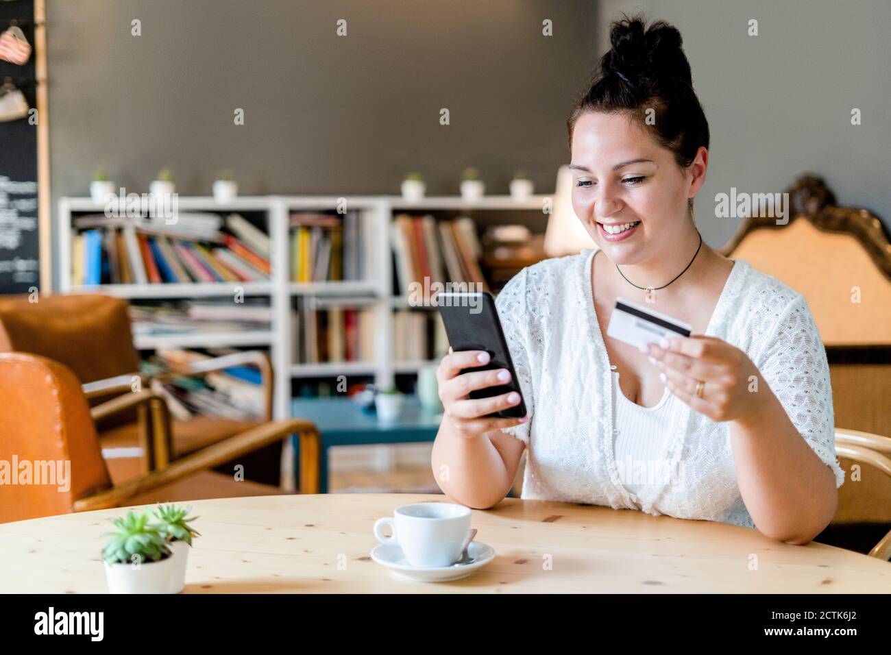 Donna sorridente con caffè sul tavolo facendo shopping online telefono cellulare in caffetteria Foto Stock