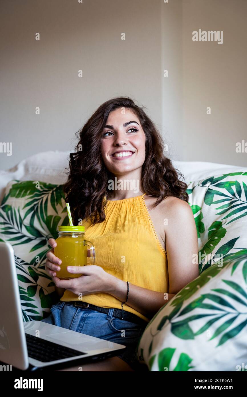 Donna sorridente premurosa che tiene il succo mentre si siede con il computer portatile acceso poltrona a casa Foto Stock