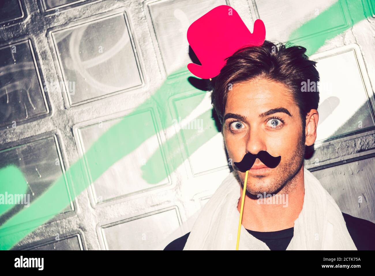 Primo piano di un uomo che indossa un falso cappello con baffi sul viso contro la parete dei graffiti di notte Foto Stock
