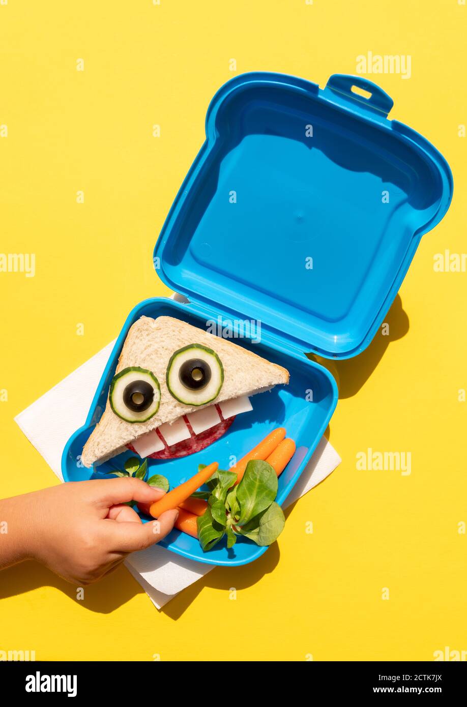 Mano della bambina che raccoglie la carota del bambino dal pranzo scatola con buffo sandwich con volto antropomorfo Foto Stock