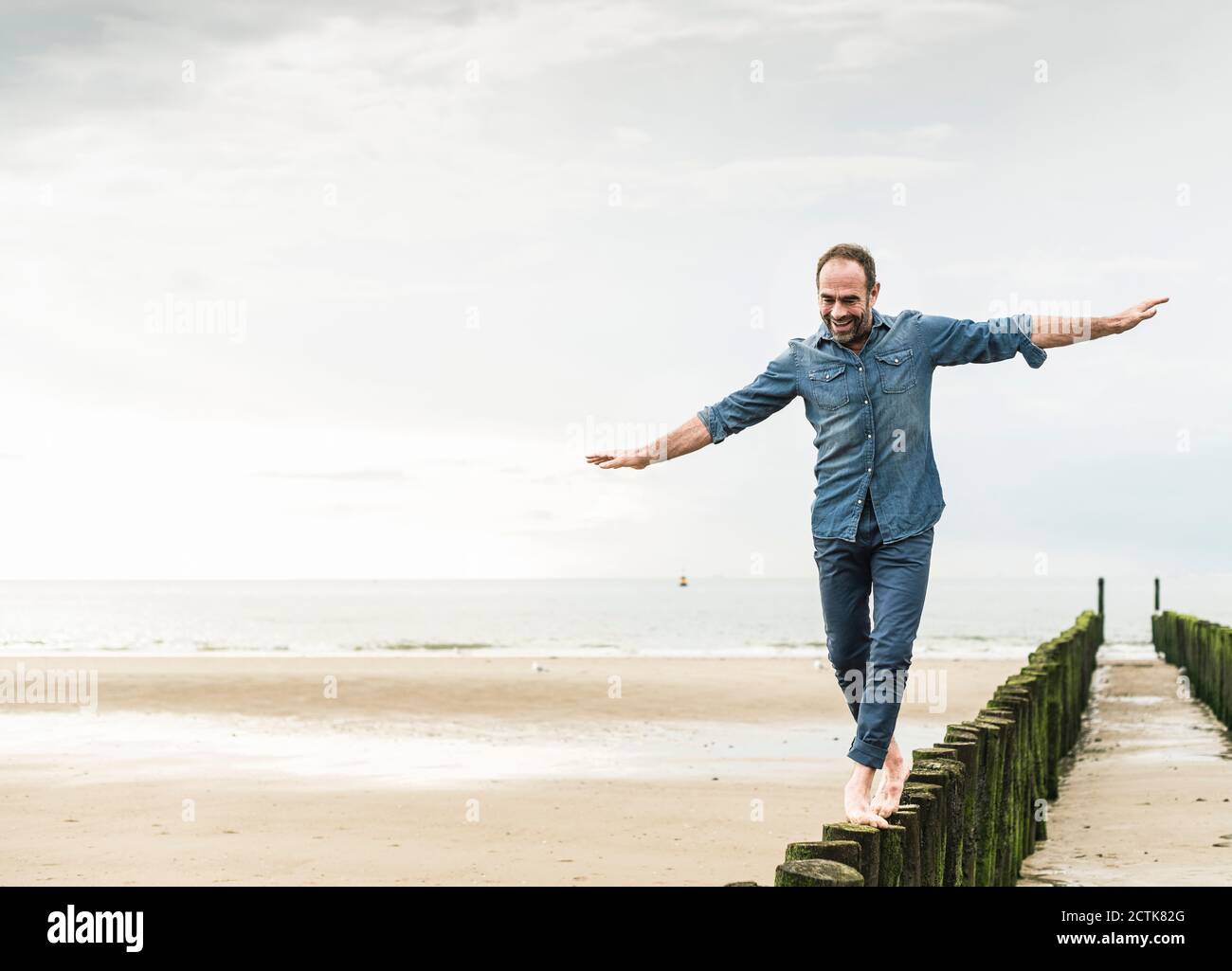 Uomo spensierato con le braccia allungate che cammina su palo di legno a. spiaggia Foto Stock