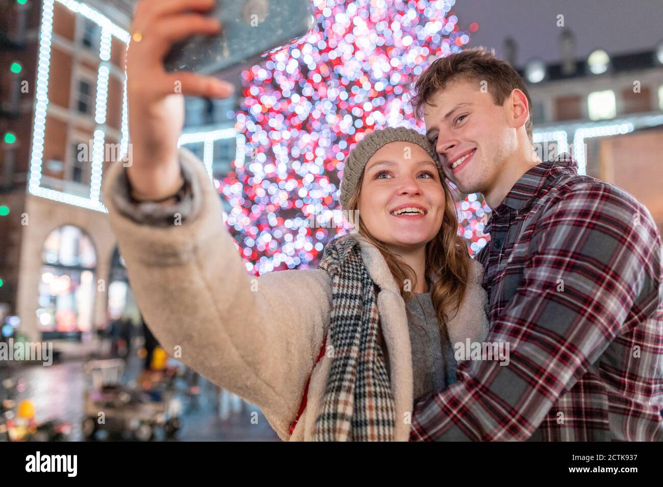 Coppia che prende selfie mentre si levano in piedi contro l'albero illuminato di Natale dentro città Foto Stock