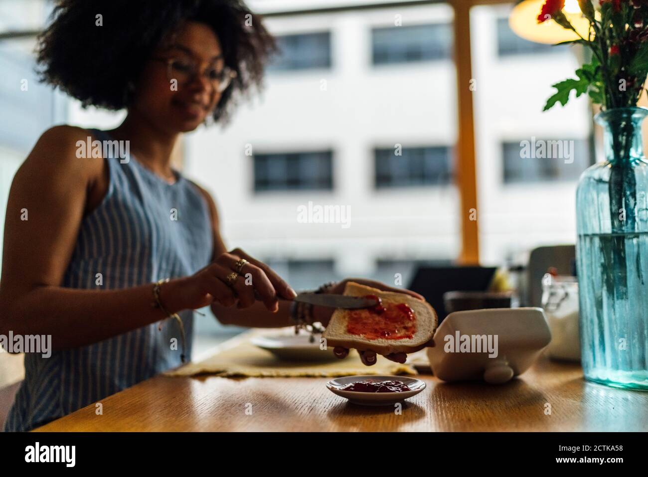 Giovane donna che spalma conserve sul pane mentre si siede a tavola Foto Stock