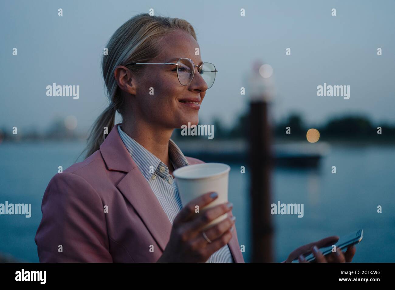 Sorridente imprenditore femminile che tiene la tazza usa e getta e il telefono cellulare mentre guardando via il crepuscolo Foto Stock