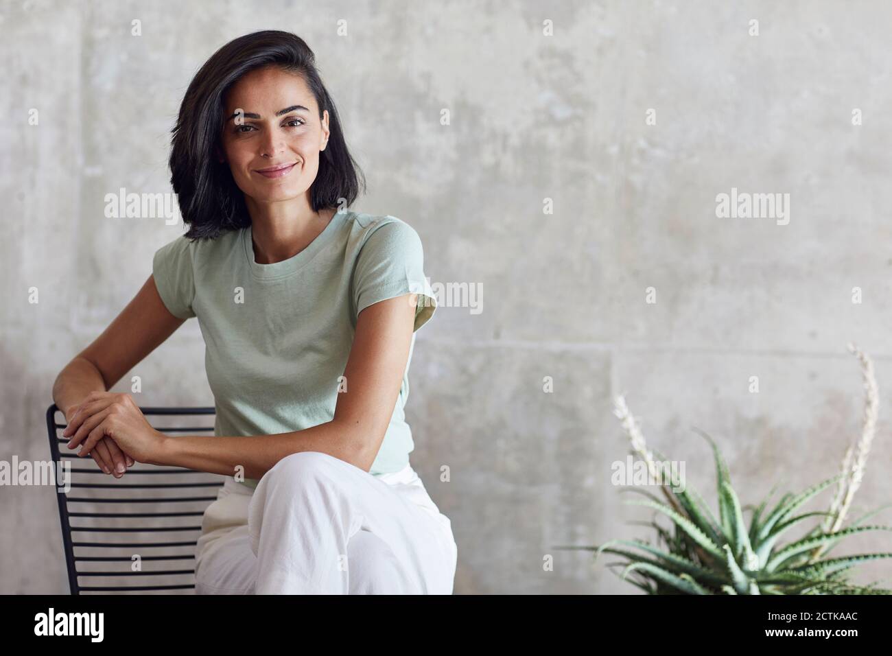 Donna d'affari sorridente seduta sulla sedia contro la parete grigia in ufficio Foto Stock