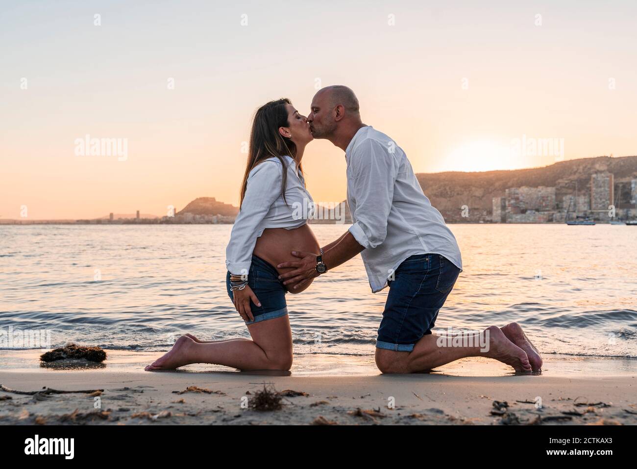 Uomo maturo baciare donna incinta mentre inginocchiandosi contro la spiaggia durante tramonto Foto Stock