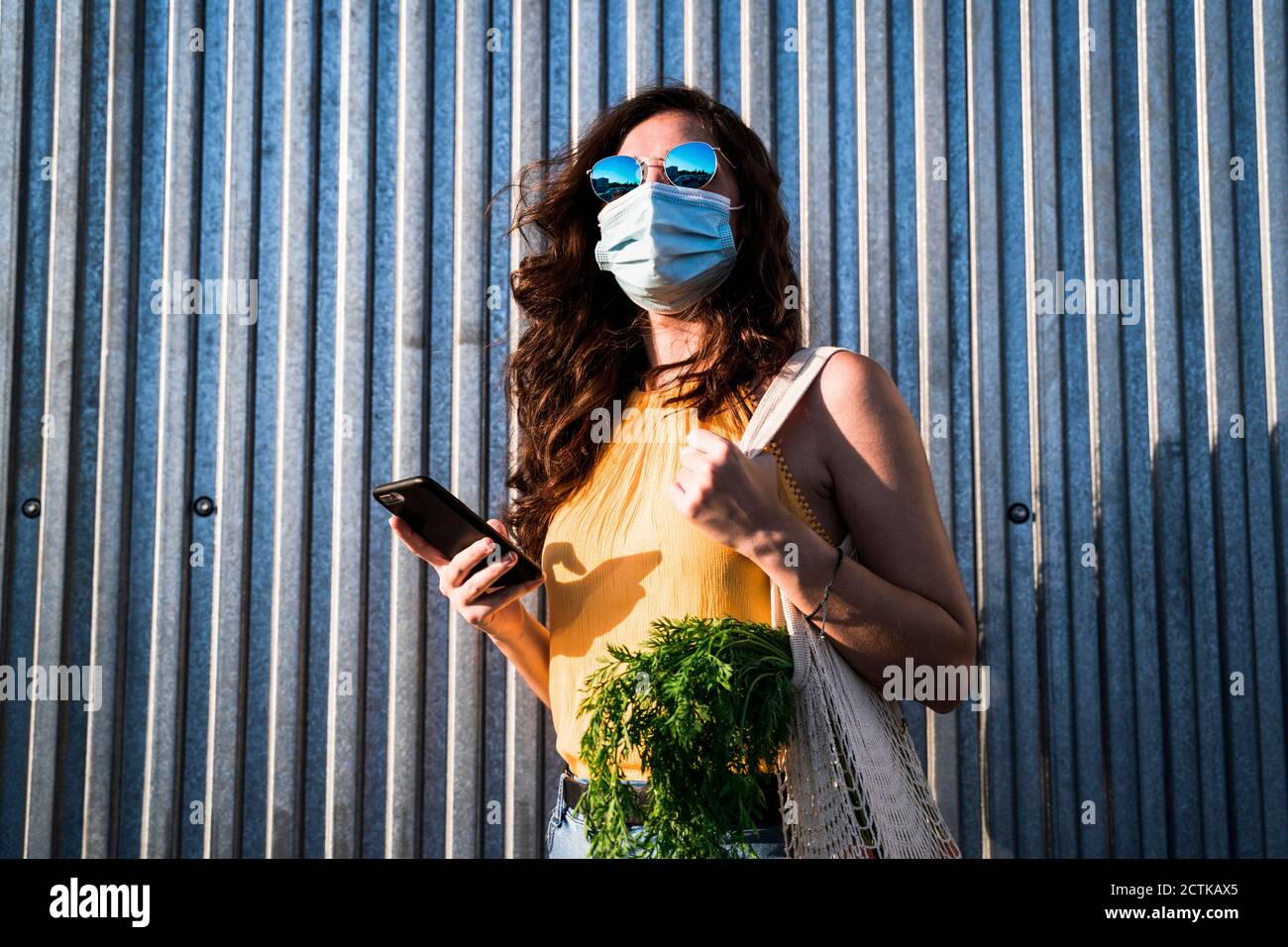 Donna che indossa la maschera mentre si è in piedi con il telefono cellulare e. sacchetto a rete riutilizzabile contro le pareti metalliche Foto Stock