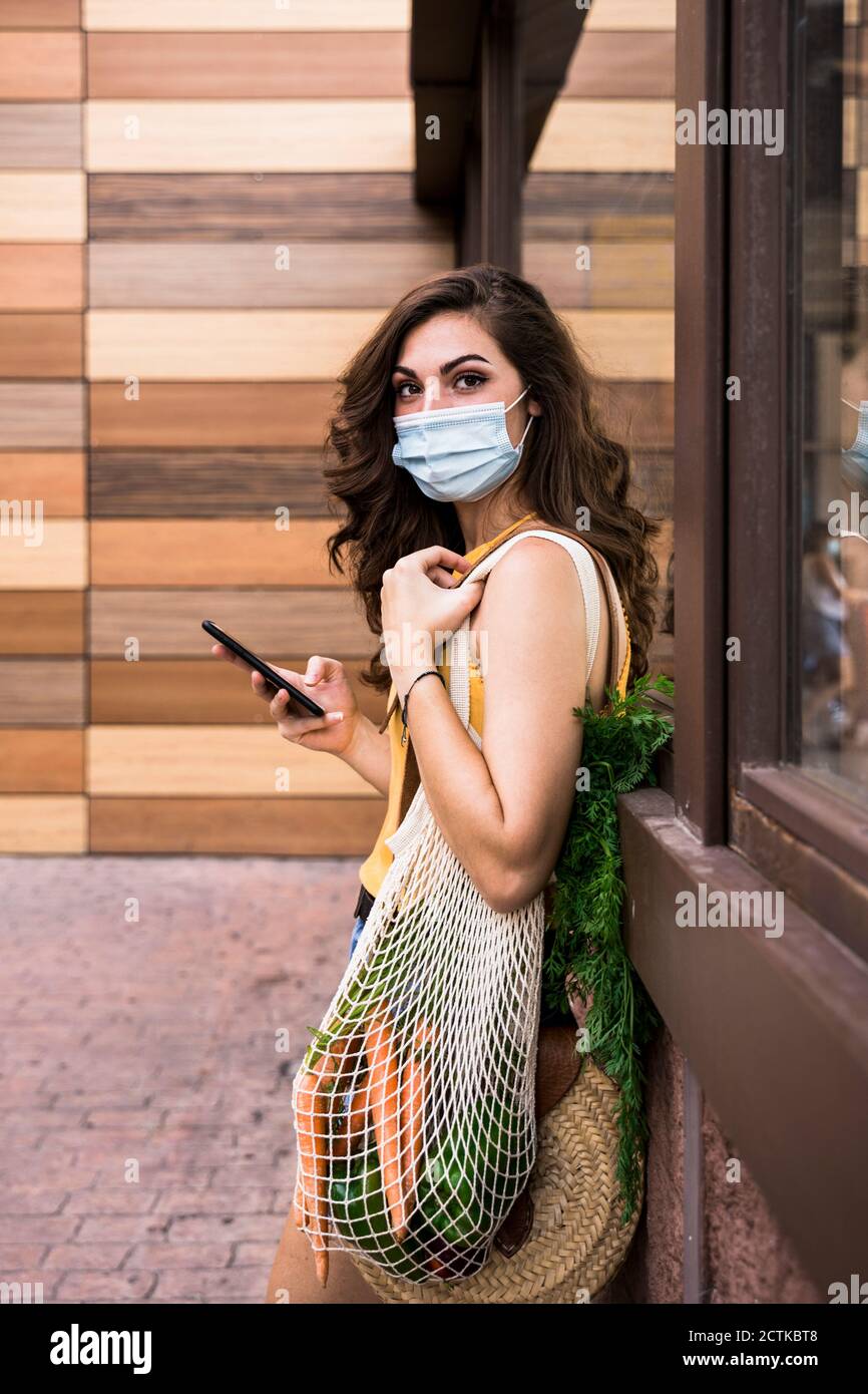 Giovane donna che indossa una maschera protettiva per il viso che tiene il telefono cellulare e. borsa a rete mentre si è in piedi contro la parete Foto Stock