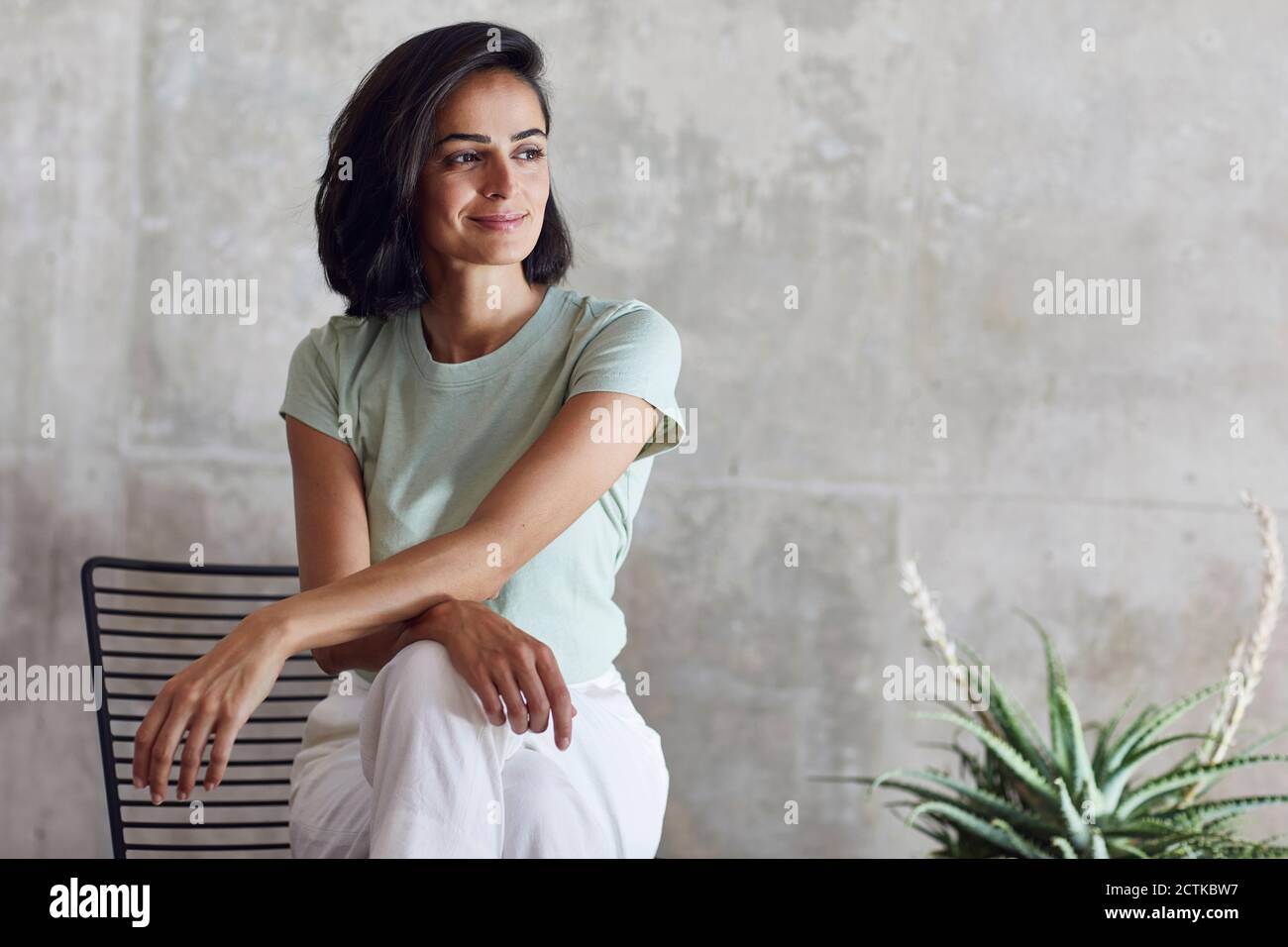 Donna d'affari premurosa sorridendo mentre si siede sulla sedia contro la parete grigia in ufficio Foto Stock