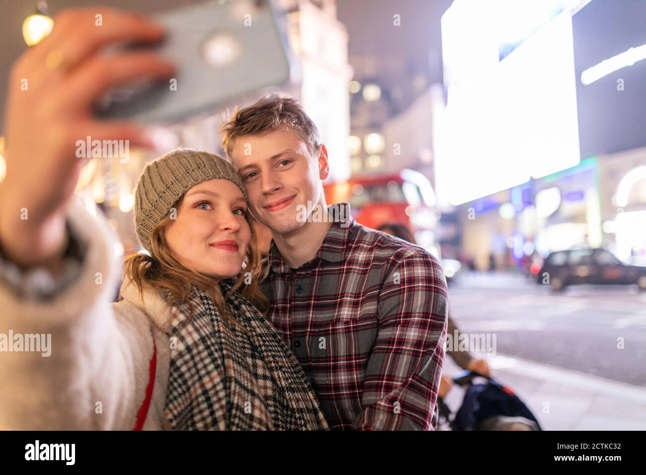 Giovane donna che prende selfie con il ragazzo mentre si trova in piedi illuminato città di notte Foto Stock