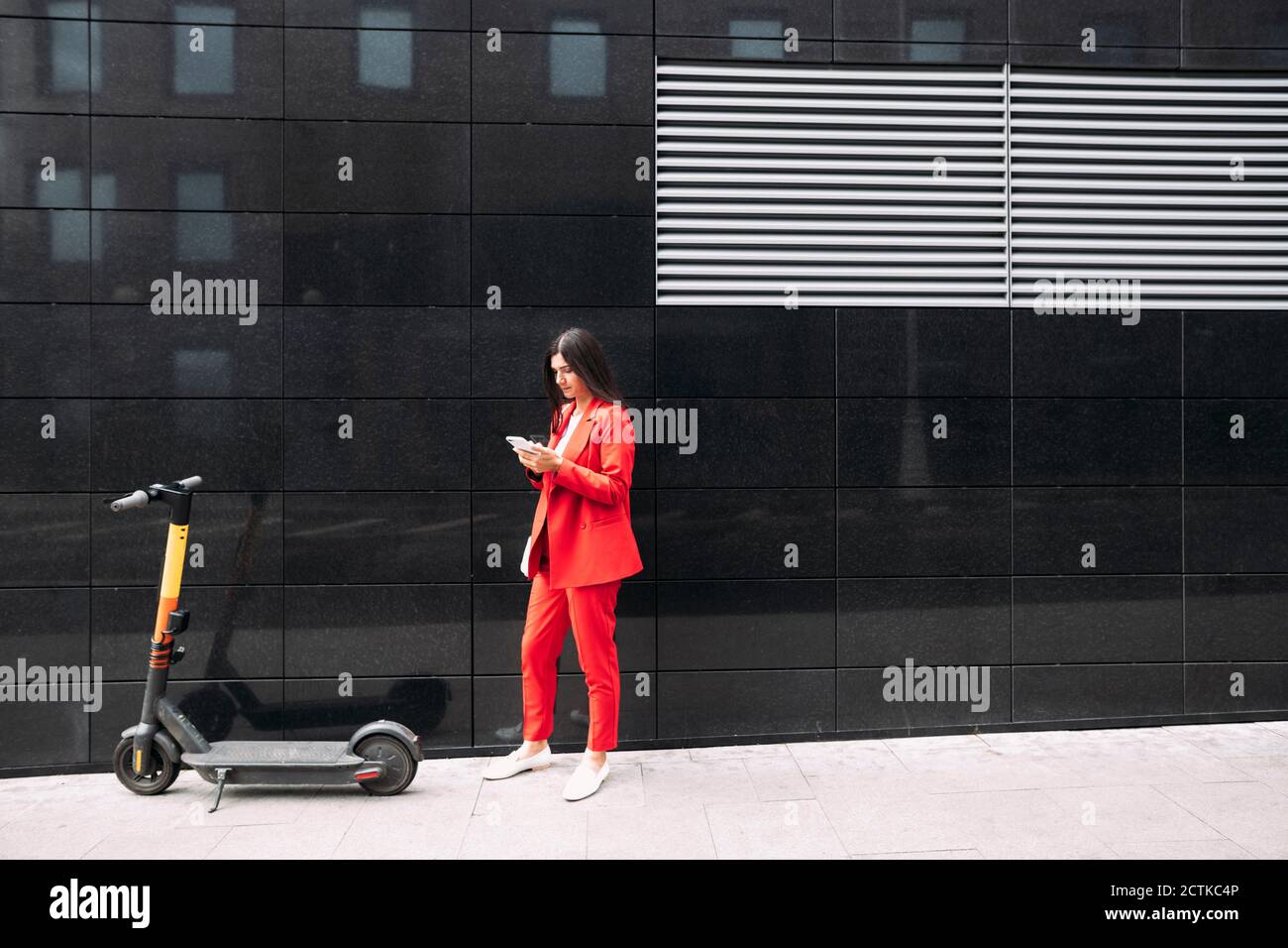 Donna imprenditore in abbigliamento da lavoro rosso con smartphone mentre si è in piedi contro l'edificio nero in città Foto Stock