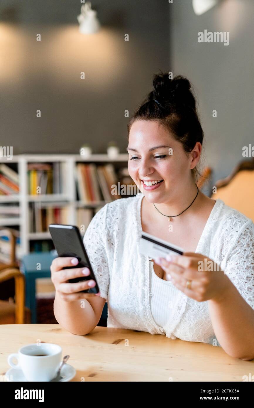 Sorridente voluttuosa donna che fa shopping in linea sopra il telefono mobile dentro caffetteria Foto Stock