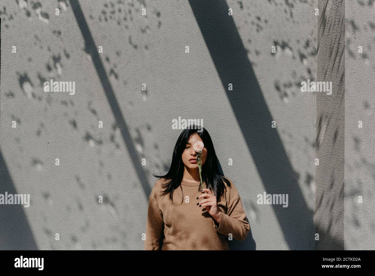 Donna che tiene il fiore davanti all'occhio mentre si sta in piedi contro muro di cemento in giornata di sole Foto Stock